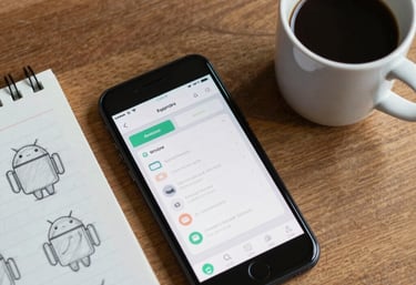 A top-down view of an Android phone and a cup of coffee on a wooden desk, with a notepad containing rough sketches of an app interface. Global / Western.