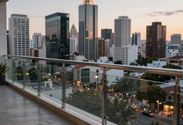A beautiful outdoor balcony featuring elegant glass railings with stainless steel posts, overlooking a modern Hispanic / Latin American city skyline at twilight.