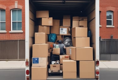 A moving truck with company branding is parked next to an open shipping container on a trailer. Two people are handling items near the container, while a car is parked in the background. The sky is overcast, and the ground appears to be asphalt.