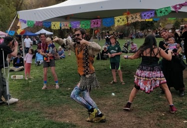 Dancers in eclectic costumes perform at an outdoor community festival under a white canopy.
