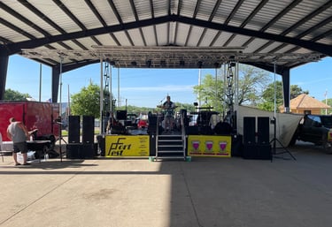 A musician performs on an outdoor stage at Fort Fest under a large metal pavilion roof with professional audio equipment.