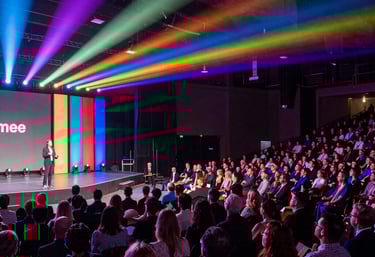 A large outdoor event with a crowd of people gathered in front of a stage. The stage is covered by a white canopy, and bright lights illuminate it. Attendees are raising their hands, suggesting enthusiasm or participation. A large screen displays an image of another part of the event, contributing to the lively atmosphere. The surrounding area is a grassy park-like setting with trees in the background.
