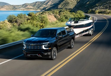 Chevrolet Silverado rebocando lancha em rodovia sinuosa à beira de lago e montanhas.