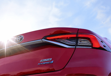 Traseira do Toyota Corolla Hybrid vermelho com iluminação solar e céu azul ao fundo em ângulo baixo.