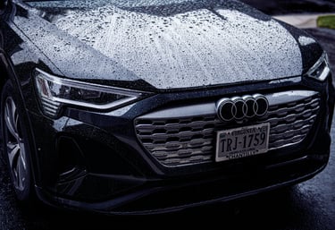 Front view of a black Audi electric vehicle covered in rain droplets with visible headlights and grille.