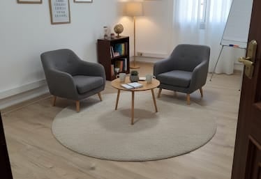 Modern therapy room with two grey armchairs, a round rug, and framed wall art on wood floors.