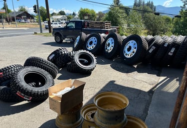 A collection of heavy-duty truck tires and chrome rims displayed on asphalt at a tire service shop.