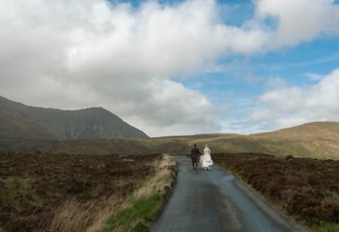 A bride and groom in a kilt walk down a road in the scenic Isle of Skye mountains.