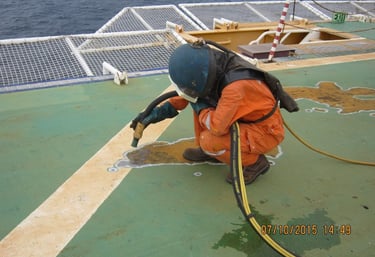 A worker in safety gear sandblasting a metal ship deck for corrosion protection and maintenance.