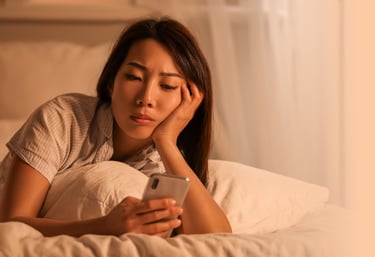 A tired woman lying in bed while using her smartphone, depicting late-night blue light exposure and insomnia