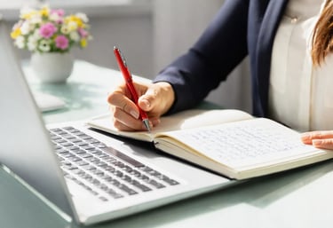 A business professional writing notes in a notebook with a red pen next to a laptop.