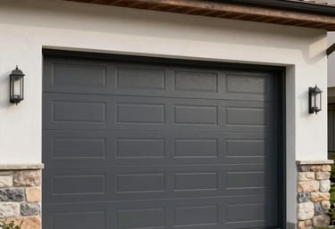A professional photograph of a modern charcoal-colored sectional garage door being operated. The house has a minimalist Southeastern European / Bulgarian design with clean stone and plaster finishes.