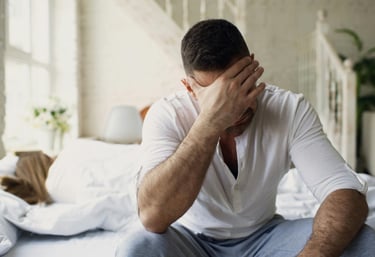Stressed man sitting on bed edge with head in hand while wife sleeps, depicting relationship problems and insomnia.