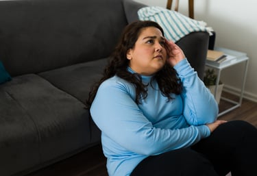 A concerned woman sitting on the floor by a sofa, holding her head to represent stress or a headache.