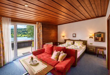 Rustic hotel room with a red sofa, wooden ceiling, and balcony overlooking a scenic lake.