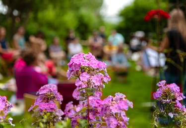 Im Vordergrund steht eine rosafarbene Blume. Im Hintergrund befindet sich eine Gruppe von Menschen, die im Garten zusammensit