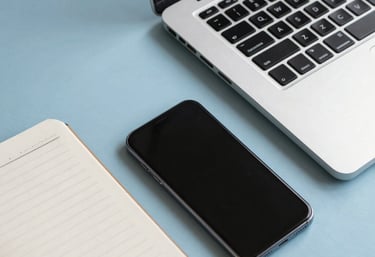 Clean, flat-lay photography of a laptop, a notebook, and a smartphone on a light blue desk. Professional and organized aesthetic.