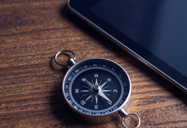 Minimalist photography of a compass and a tablet on a wooden desk. Deep navy blue shadows and warm light, symbolizing direction and strategy.