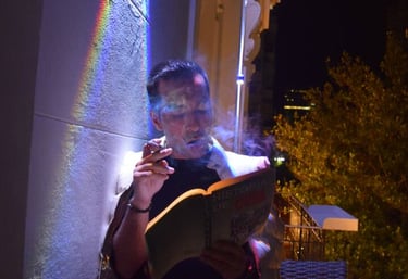 A man smoking a cigar and reading a book on a balcony at night under a rainbow light effect.
