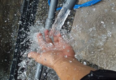 Fresh water splashing onto a person's hand from a grey PVC pipe outdoors.