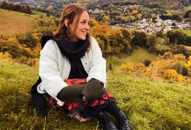 Smiling woman sitting on a grassy hill overlooking a scenic autumn valley with fall foliage and a small village.