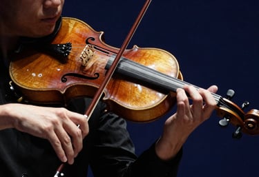 Close-up photography of a violinist's hands and bow during a performance. Dramatic lighting against a dark navy blue background highlights the rich wood tones.
