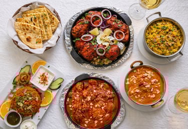 An Indian food spread featuring butter chicken, tandoori chicken, garlic naan, and biryani rice on a white tablecloth.