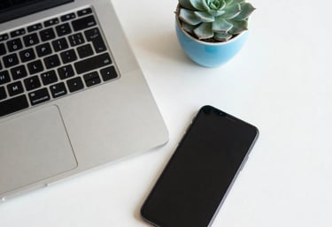An overhead shot of a clean, minimalist desk with a silver laptop and a smartphone. A small cyan blue succulent pot adds a touch of life to the off-white surface.