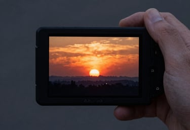 A hand-held professional video monitor showing a vibrant sunset scene being filmed. Dark grays and warm oranges.