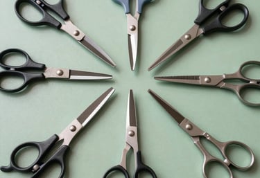 An overhead flat lay photograph of various specialized scissors arranged neatly on a muted green surface. Includes fabric shears, hair styling scissors, and precision craft tools. The setting is a bright, modern North American / US workshop with clean, even lighting.