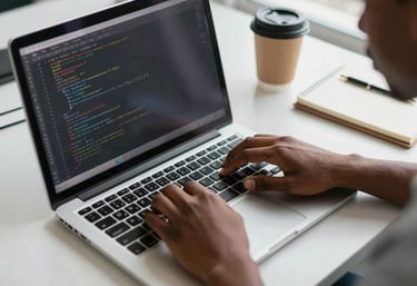 Close-up overhead shot of a young African developer's hands on a laptop keyboard in a bright tech lab, lines of code visible on screen, natural daylight from a side window, a coffee cup and notebook nearby