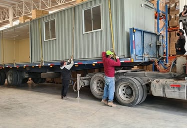 Logistics workers securing a modular portable shipping container office onto a semi-truck trailer for transport.