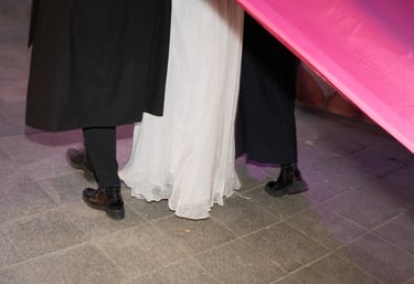 Elegant guests in formal attire and black boots standing near a white dress and bright pink fabric banner.