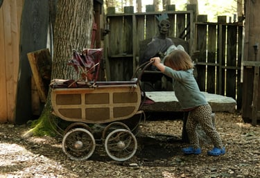 A toddler pushes a vintage stroller in a spooky forest setting near a skeletal king prop.