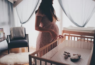 A pregnant woman stands in a sunny nursery, holding her baby bump next to a wooden crib.