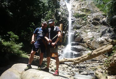 Two survival participants are standing in front of a waterfall holding a machete during the trip