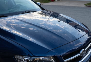A close-up of a glossy, freshly washed car reflecting a clear blue sky in a North American / US driveway. Water beads sit perfectly on the hood. Colors: Steel Blue and Dark Navy Blue.