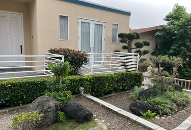 Modern Japanese zen garden with a stone lantern, manicured shrubs, and lava rocks in front of a tan house.