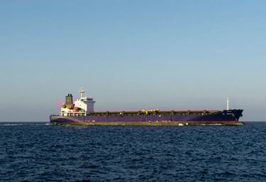A wide-angle shot of a cargo freighter ship leaving the harbor into the deep navy open sea under a clear sky blue morning sky.