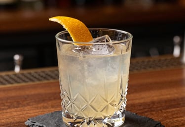A sophisticated cocktail in a crystal glass on a dark slate coaster. The drink is garnished with a thin citrus peel. Warm bronze lighting, South American high-end bar setting, blurred background of wood and glass.