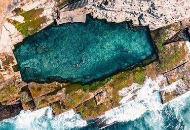 Aerial view of the Bogey Hole in Newcastle with swimmers, rocky coastline, and clear ocean water.