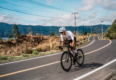 A cyclist in white gear riding a black road bike on a winding mountain highway.