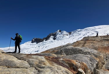 A hiker with trekking poles stands on a rocky ridge overlooking a snow-covered mountain glacier.