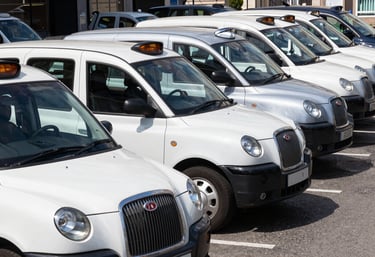 A row of clean, professional white and silver taxis parked neatly at a European / British transit hub. Clear, bright daylight, emphasizing organization and readiness.