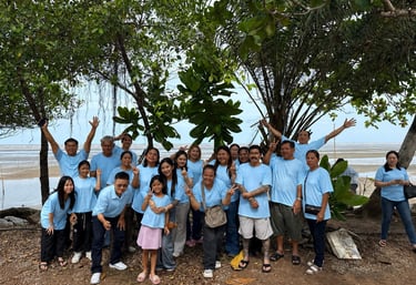 A large group of people in matching light blue t-shirts posing for a photo outdoors near a beach with trees.