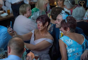Wedding guests in elegant formal attire smiling and taking a group selfie with a smartphone at a reception table.