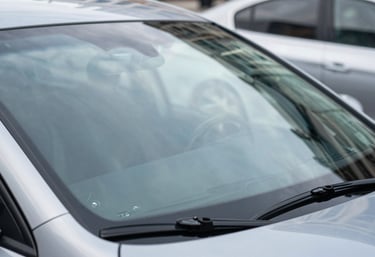 A close-up of a perfectly installed, clear windshield on a modern car, reflecting a bright North American / US city environment. The image uses steel blue and pale mist grey colors to convey quality and clarity.