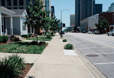 A bright daytime view of a North American / US city street in Detroit, featuring clean sidewalks and well-maintained property landscaping in a medium blue tone.