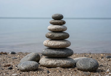 A macro photograph of smooth, rounded river stones stacked in a small cairn on a sandy North American shore. The water is calm and reflecting a light blue-grey sky, conveying balance and serenity.