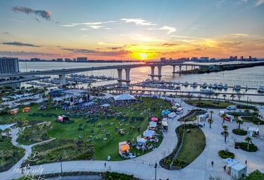 Aerial view of a waterfront park festival with a sunset backdrop over a bridge and marina.